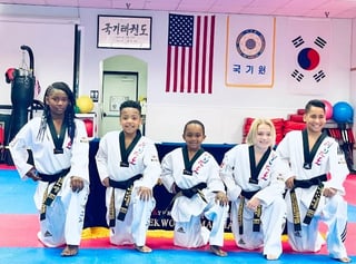 Five young taekwondo athletes in white uniforms with black belts posing together on a mat in a training facility with flags on the wall