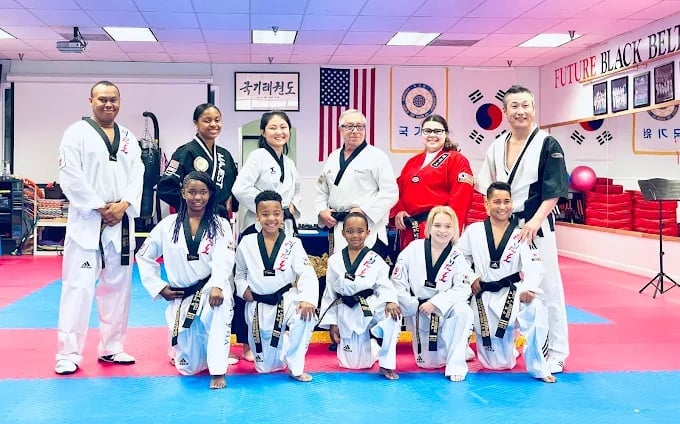 Taekwondo instructors and students in uniforms posing together on a mat in a dojo with Korean flags